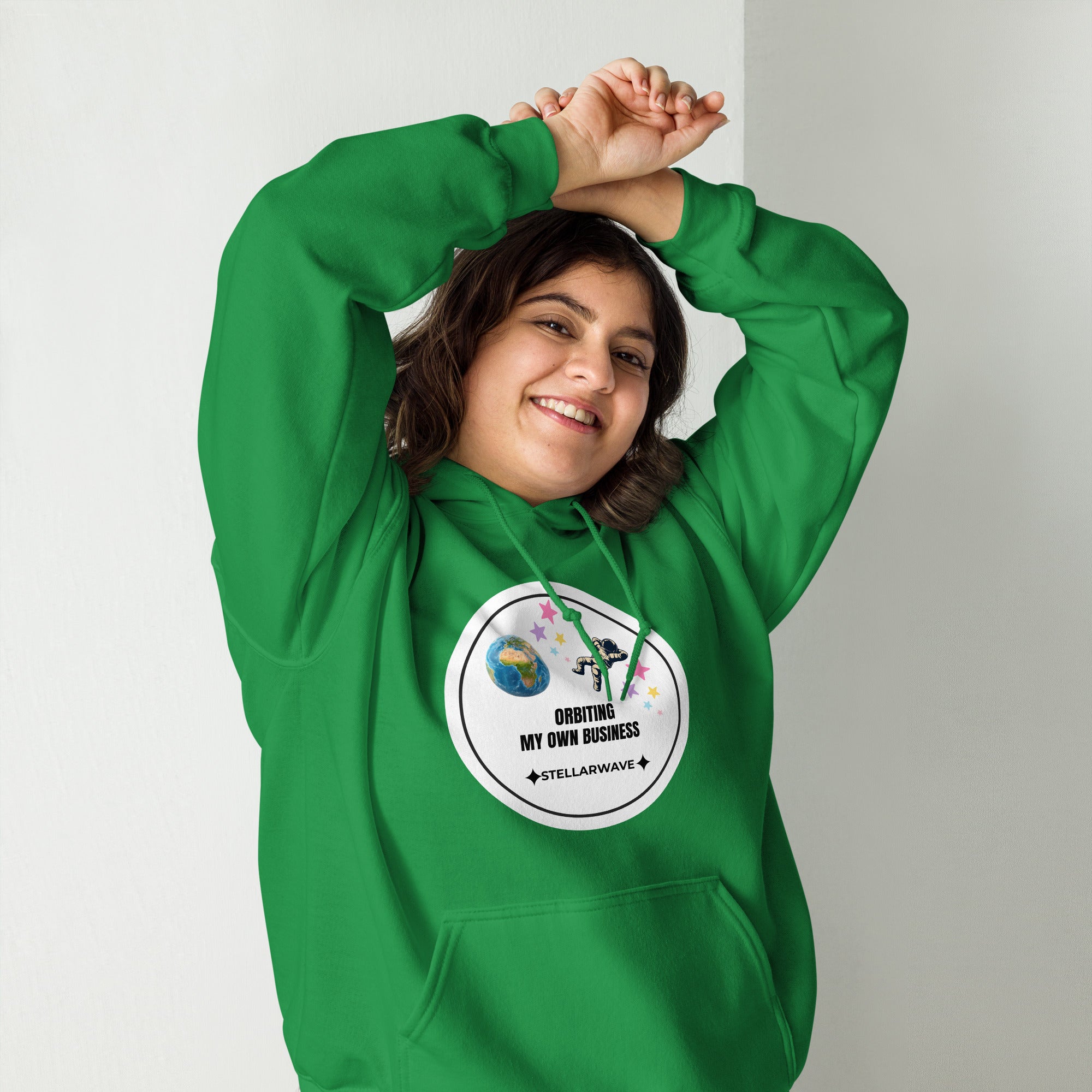 Woman wearing a green Cosmos Comfort Collection - Orbiting My Own Business - Unisex Hoodie with space-themed graphic and motivational text, smiling with arms raised.
